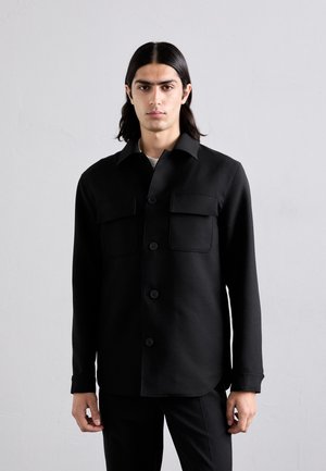 Schwarze Blusenjacke mit Kragen, zwei vorderen Taschen und langen Ärmeln, aus einem glatten Stoff mit matter Oberfläche gefertigt.