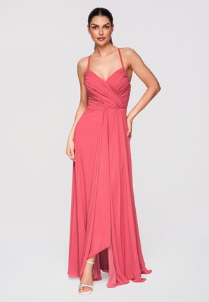 Femme en robe corail rose longue jusqu’au sol, à fines bretelles spaghetti et corsage froncé, debout, une main sur la hanche, sur fond uni.