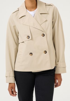 Trench-coat beige croisé avec des boutons foncés, de larges revers et des brides aux poignets, porté par-dessus une chemise blanche et un pantalon noir.
