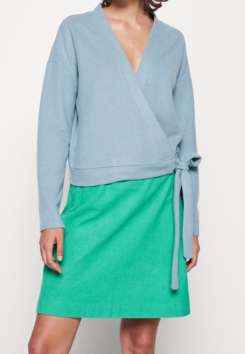 Femme portant un pull cache-cœur bleu clair avec un lien latéral sur une jupe vert vif longueur genou, sur un fond uni.