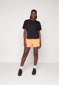 Zwarte katoenen T-shirt in combinatie met feloranje shorts. Model draagt zwarte sportschoenen met witte sokken en staat tegen een neutrale achtergrond.