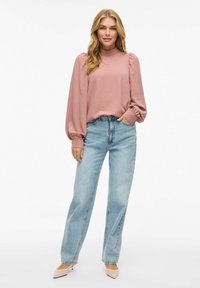 Kvinna klädd i en långärmad dammig rosa blus med puffärmar, ljusblå högmidjade jeans och beige spetsiga klackar, står avslappnat.