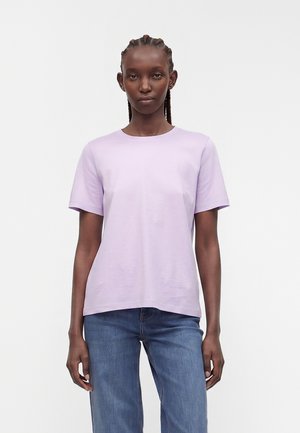 Femme aux cheveux tressés portant un t-shirt à manches courtes violet clair et un jean bleu sur un fond blanc uni.
