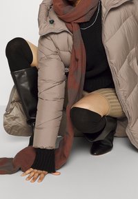 Person der sidder på hug iført en beige puffjakke, sorte støvler til knæene, sorte knæstrømper, et brunt t�ørklæde og en tan strikket nederdel.