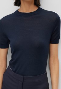 Mörkblå kortärmad stickad topp med rundad halsringning; mjuk textur, något genomskinlig, matchad med mörka skräddarsydda byxor.