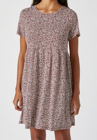 Vrouw in een jurk met korte mouwen en knielange lengte, met een bruine en witte bloemenpatroon en een geraffineerde taille, staand tegen een effen achtergrond.