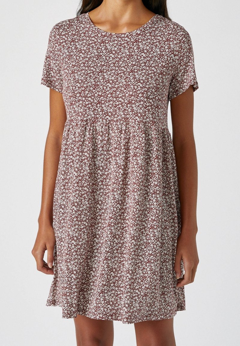 Vrouw in een jurk met korte mouwen en knielange lengte, met een bruine en witte bloemenpatroon en een geraffineerde taille, staand tegen een effen achtergrond.
