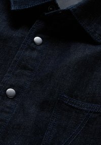 Camicia in denim blu scuro con collo classico, bottoni a pressione argentati e una tasca sul petto con dettagli di cuciture a contrasto.