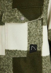 Gros plan sur la poche d'une veste en tissu à carreaux verts et blancs, avec un rabat fixé par un bouton-pression argenté et un petit patch noir avec un motif blanc.