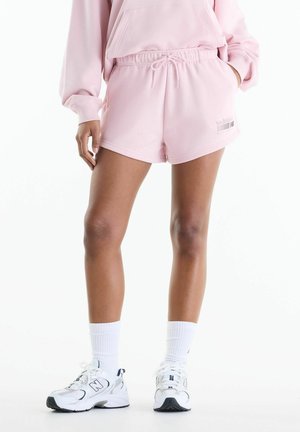Person iført lyserøde sweatshorts og matchende hættetrøje med hvide crew-sokker og hvide New Balance-sneakers, der står mod en hvid baggrund.