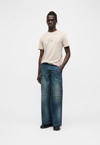 Άνδρας φορώντας μπεζ γραφικό t-shirt, φαρδιά μπλε τζιν και μαύρα παπούτσια, στέκεται με το ένα χέρι στην τσέπη σε λευκό απλό φόντο.