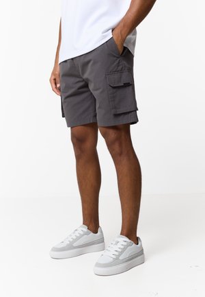 Mężczyzna w ciemnoszarych szortach cargo i białych sportowych butach, stojący z jedną ręką w kieszeni na jednolitym białym tle.