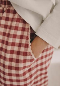 Rote und cremefarbene Gingham-Hosen mit geschnittenem Taschenrand und einem teilweise sichtbaren weißen Langarmoberteil. Aus Baumwollmaterial.