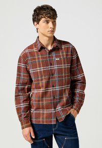 Camicia a quadri marrone con bottoni, caratterizzata da strisce blu, gialle e bianche, realizzata in un tessuto testurizzato, con una tasca sul petto visibile e colletto.