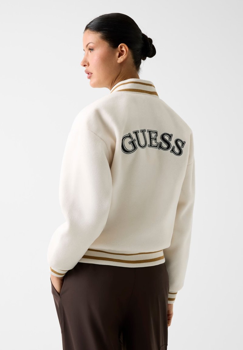 Veste bomber en polaire blanche avec col et poignets côtelés à rayures dorées, ornée du logo "GUESS" noir brodé dans le dos.