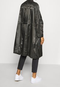 Person med en glänsande svart lång regnkappa, avkortade mörka jeans och vita sneakers, som står mot en enhetlig ljus bakgrund.