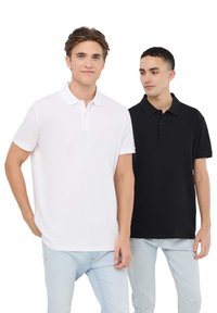 Due polo shirt: uno bianco con colletto classico e maniche corte, l'altro nero, entrambi in cotone. Indossati con jeans azzurri chiari.