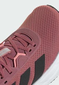 Light pink athletic shoe with a textured mesh upper, contrasting black stripes, and flexible rubber sole. Features flat laces and padded collar.