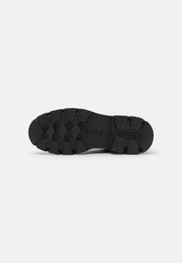 Semelle de chaussure en caoutchouc noir avec des motifs de bande de roulement profonds pour l'adhérence. Présente un logo de marque le long de l'arche. Surface lisse avec bords texturés.