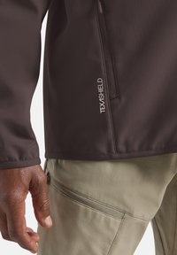 Veste softshell brune avec une poche zippée et un logo TEXASHIELD ; assortie à un pantalon kaki avec une poche latérale zippée.