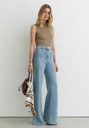 Kvinde med blondt hår iført brun ærmeløs top og blå vidde højtaljede bredbenede jeans, holder brun håndtaske med mønstret tørklæde.