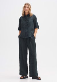 Camicia in lino verde scuro con maniche corte, due tasche frontali, abbinata a pantaloni larghi e morbidi della stessa tonalità e sandali flat.