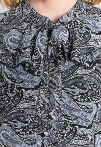 Blusa preta com padrão paisley em tons de cinza e azul. Apresenta um decote franzido e detalhe de laço, com tecido macio e texturizado.