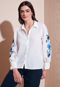 Blouse en lin blanc avec broderies florales bleues sur les manches, fermeture à boutons sur le devant et col classique ; possède des manches flottantes et froncées.