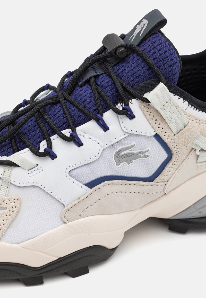 Sneakers Lacoste con una combinazione di rete e camoscio bianco, grigio e blu navy, con lacci neri e logo coccodrillo grigio. Suola in gomma testurizzata.