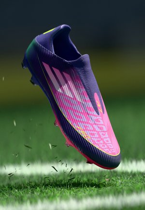 Lila és rózsaszín foci cipő, texturált felsőrésszel, ferde formával és neon sárga részletekkel, fű fölé emelve, látható a cleat és a csíkok.