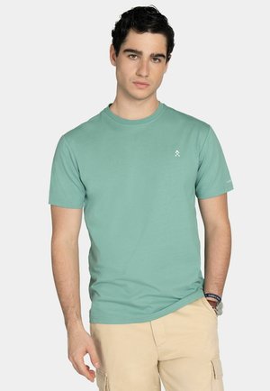 Jeune homme aux cheveux bouclés foncés portant un t-shirt à manches courtes bleu sarcelle clair et un short cargo beige, debout avec une main dans la poche.