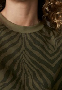 Zbliżenie osoby w oliwkowozielonej koszuli w paski w zebrę, z widocznymi kręconymi włosami po prawej stronie.