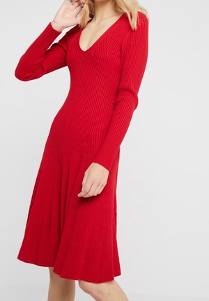 Femme portant une robe en maille côtelée rouge à manches longues et col en V, debout devant un fond clair uni.