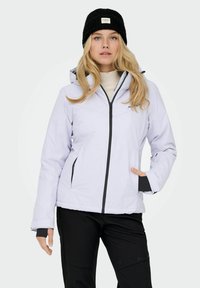 Ženska nosi belo zip-up kapuco, črne hlače, črno kapo in bež pulover z visokim ovratnikom, stoji z eno roko v žepu.