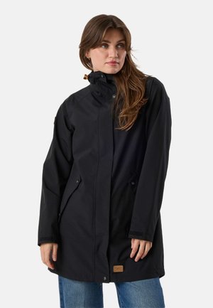 Vrouw draagt een lange zwarte regenjas met hoge kraag en drukknoopsluiting, gecombineerd met een blauwe spijkerbroek, staand tegen een effen achtergrond.