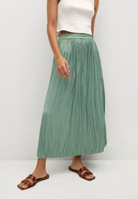 Vrouw in een witte mouwloze crop top, groene geplooide maxi rok en bruine platte sandalen, staand voor een effen achtergrond.