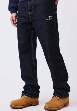 Homme portant un jean bleu foncé avec des coutures blanches et des bottes de travail marron, jean avec un petit logo blanc "PXP" sur la cuisse.