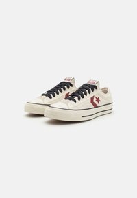 Baskets en toile crème avec lacets noirs, logo étoile rouge, semelle en caoutchouc et rayures noires contrastantes le long de la base. Design sportif classique.