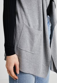 Cardigan grigio a maglia con tasca frontale, abbinato a una maglietta nera a maniche lunghe a costine, indossato sopra jeans azzurri chiari.
