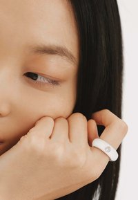 Anillo de resina blanca con una textura suave, que presenta un pequeño acento de gema clara. La mano del modelo descansa contra la cara, mostrando el anillo.