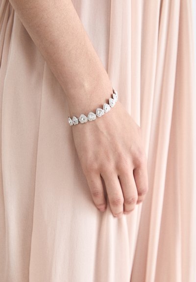 Bracelet en argent avec des pierres précieuses en forme de poire et circulaires alternées, montées en griffes, porté sur une main contre un tissu doux et léger.