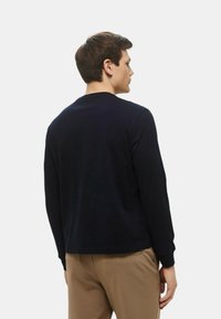 Camicia a maniche lunghe di colore blu scuro con scollo rotondo, texture liscia e vestibilità comoda. Presenta polsini a coste e un design sul retro. I pantaloni sono di colore beige.