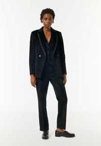 Schwarzer maßgeschneiderter Anzug aus softem Material, bestehend aus einem doppelt geknöpften Blazer und passenden Hosen, ergänzt durch eine schwarze Weste und Loafer.