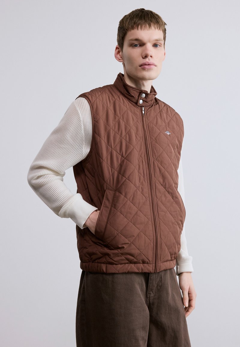 Brown quilted vest with a high collar, side pockets, and zipper closure, layered over a white knit long-sleeve shirt. Smooth texture.