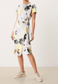 Robe à manches courtes en tissu doux avec une impression abstraite multicolore dans des teintes de jaune, noir et pastel, attachée à la taille. Baskets beiges.