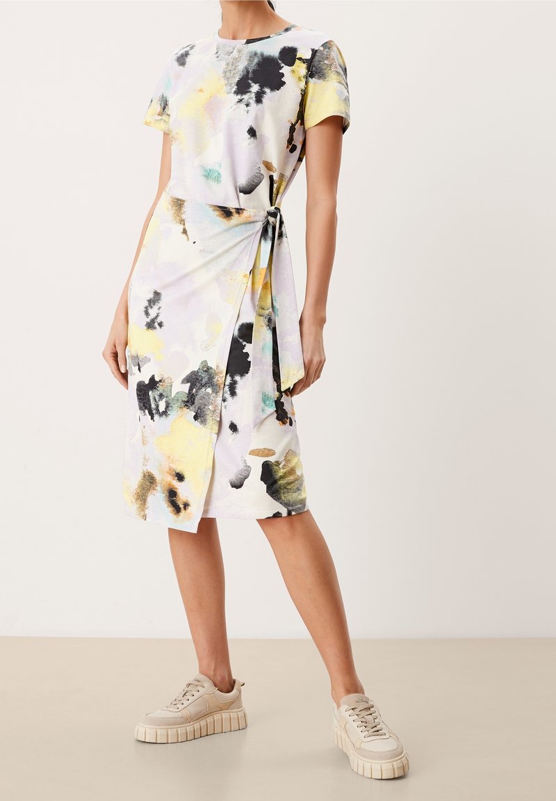 Robe à manches courtes en tissu doux avec une impression abstraite multicolore dans des teintes de jaune, noir et pastel, attachée à la taille. Baskets beiges.