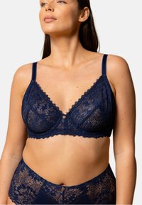 Soutien-gorge en dentelle bleue marine avec un délicat motif floral, bonnets doux, bords festonnés et bretelles ajustables. Accent en bouton argenté au centre.