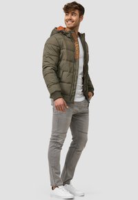 Oliven grøn dunjakke med hætte, der har en orange indvendig foring og logodetalje. Sammen med lysegrå jeans og hvide sneakers.