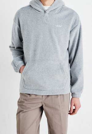 Hombre con sudadera con capucha tipo pulóver de Levi's color gris claro con bolsillo frontal y pantalones beige, de pie frente a un fondo blanco liso.