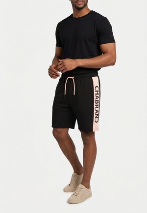 Hombre de pie con camiseta negra, pantalones cortos negros con paneles laterales rosas y texto "CHABRAND", y zapatillas beige sobre fondo blanco.